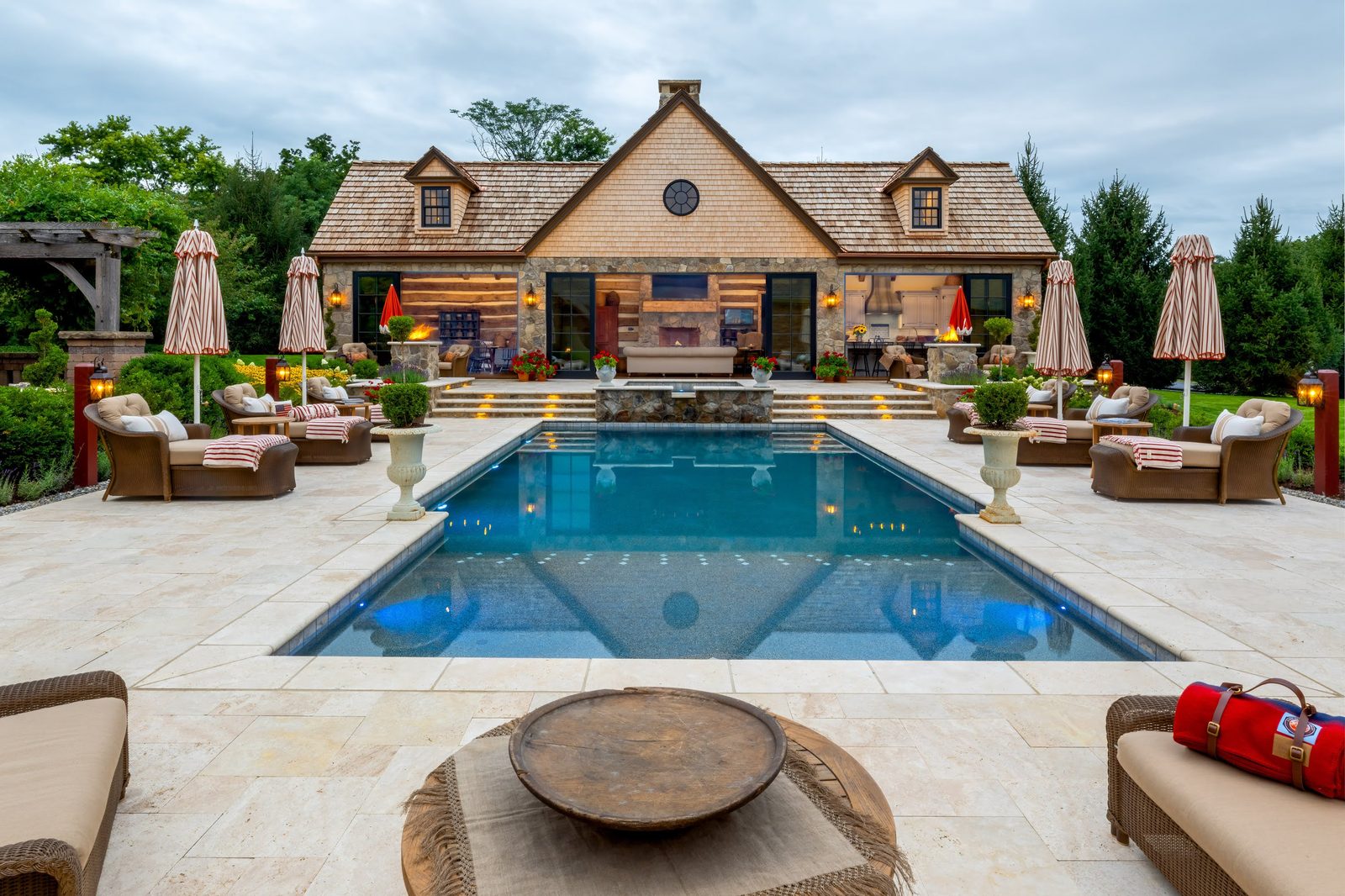 Symmetrical evening view of the pool house with stone, cedar shake, and warmly lit interior